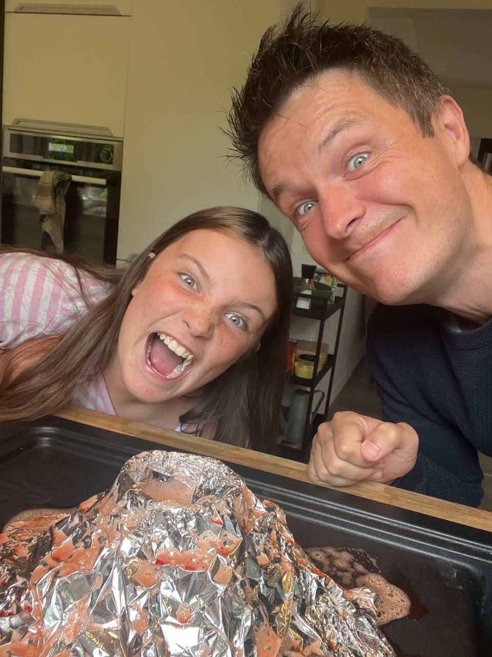 A father and his daughter grinning over a foil volcano erupting on a baking tray